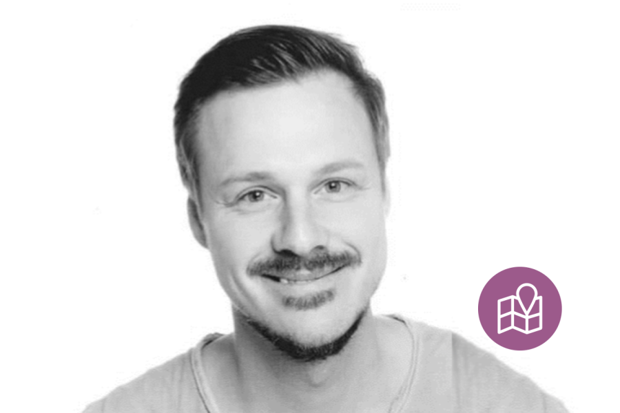 Smiling man in a grayscale head-and-shoulders portrait with short dark hair; a purple location pin badge appears on the lower-right.